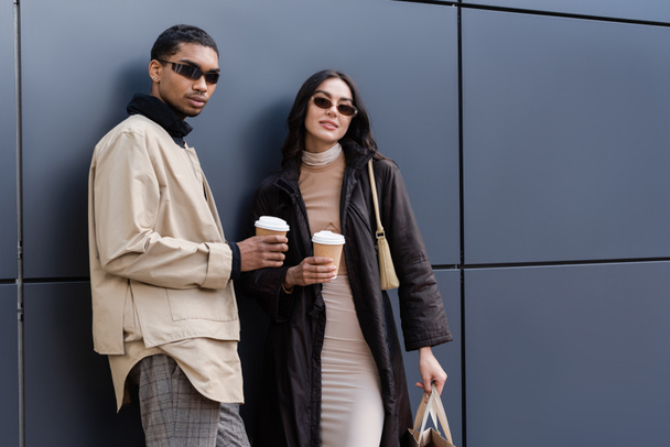 hombre afroamericano y mujer joven en gafas de sol con estilo sosteniendo vasos de papel y bolsa de compras - Foto, Imagen