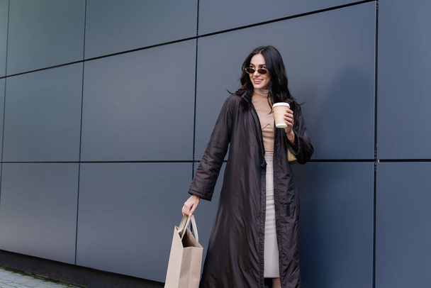 cheerful young woman in stylish outfit and sunglasses holding paper cup and shopping bag near building  - Zdjęcie, obraz