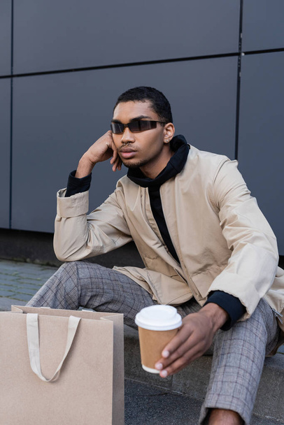 african american man in sunglasses and autumnal outfit sitting with coffee to go near shopping bag  - Foto, Imagen