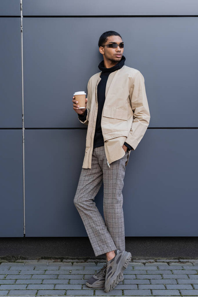 stylish african american man in stylish autumnal outfit holding paper cup and standing with hand in pocket - 写真・画像