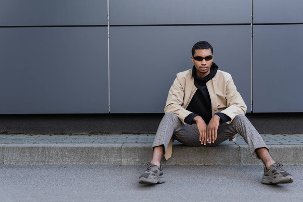 full length of young african american man in autumnal outfit and stylish sunglasses sitting near building - Photo, Image