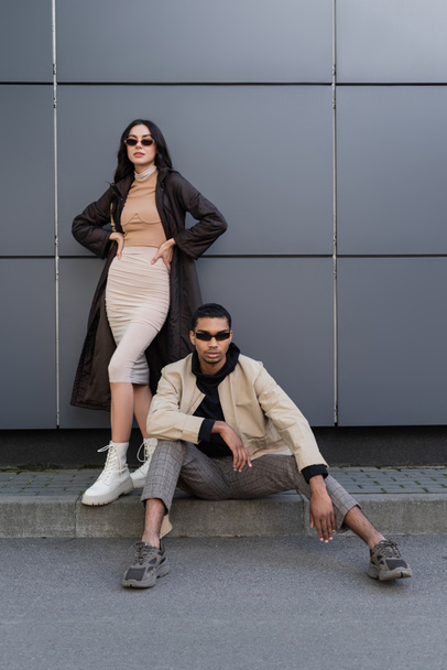 full length of stylish multiethnic couple in trendy autumnal outfits and sunglasses posing near building  - Photo, Image