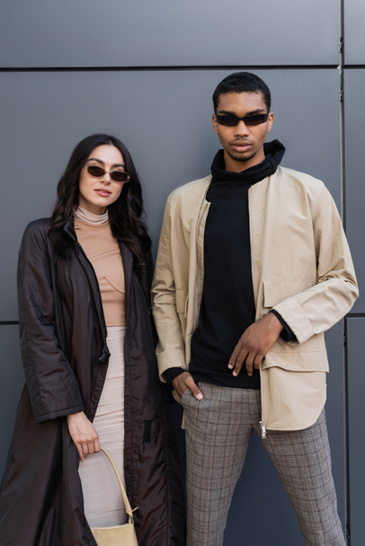 young multiethnic couple in trendy autumnal outfits and sunglasses posing near building  - Foto, Bild