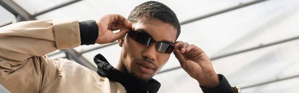low angle view of young african american adjusting stylish sunglasses under roof, banner - Фото, изображение