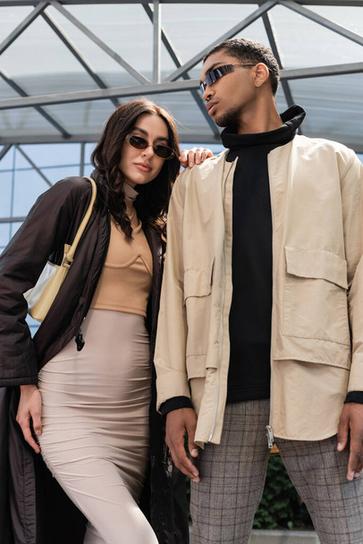 low angle view of stylish interracial couple in sunglasses and stylish outfits looking at camera  - Foto, Bild