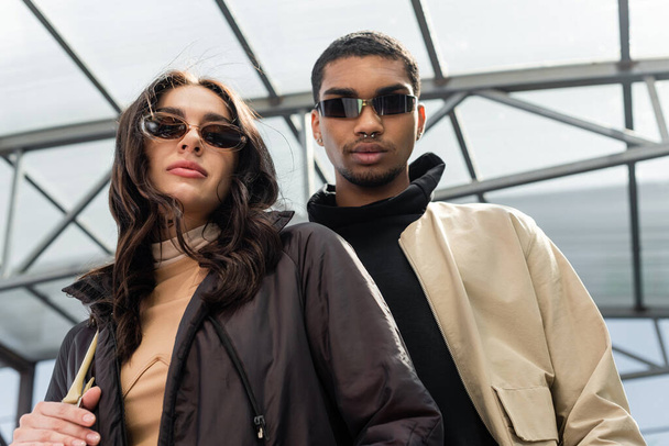 low angle view of stylish multiethnic couple in sunglasses looking at camera  - Φωτογραφία, εικόνα