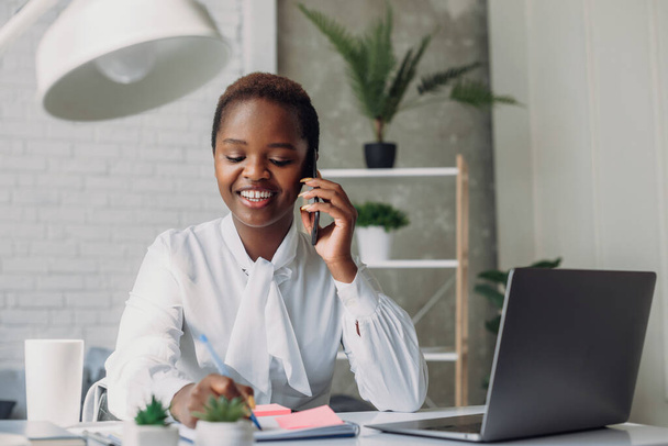 Lächelnde afrikanisch-amerikanische Geschäftsfrau, die mit dem Handy telefoniert und sich Notizen in ihrem Notizbuch macht, sitzt an ihrem Arbeitsplatz im Büro. Kleinunternehmen - Foto, Bild
