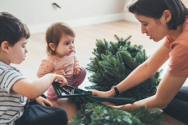 Η μητέρα και τα δύο παιδιά της συναρμολογούν το τεχνητό χριστουγεννιάτικο δέντρο, περνώντας το Σαββατοκύριακο στολίζοντας το σπίτι για τις γιορτές. Παραδοχές για μια οικογένεια - Φωτογραφία, εικόνα