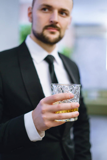 Ένας νεαρός με γενειάδα, ντυμένος με επαγγελματικό κοστούμι, κρατώντας ένα ποτήρι ουίσκι. Επιχειρηματίας που πίνει αλκοόλ σε εστιατόριο. - Φωτογραφία, εικόνα