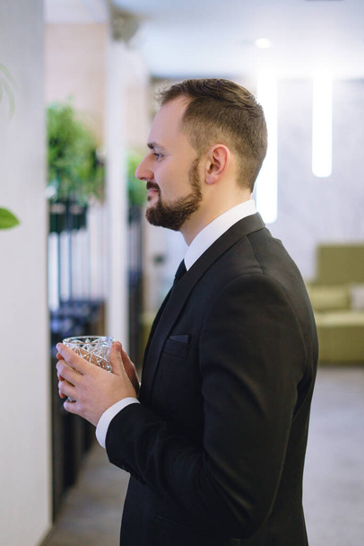 Mladý muž s vousy, oblečený v obchodním obleku, držící sklenici whisky. Podnikatel pije alkohol v restauraci - Fotografie, Obrázek