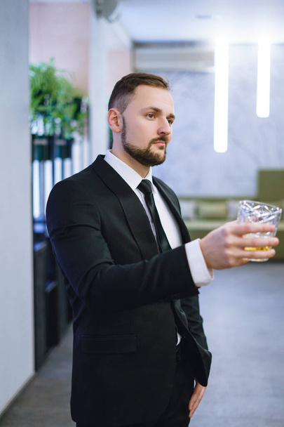 A young man with a beard, dressed in a business suit, holding a glass of whiskey. A businessman drinking alcohol in a restaurant - Фото, изображение