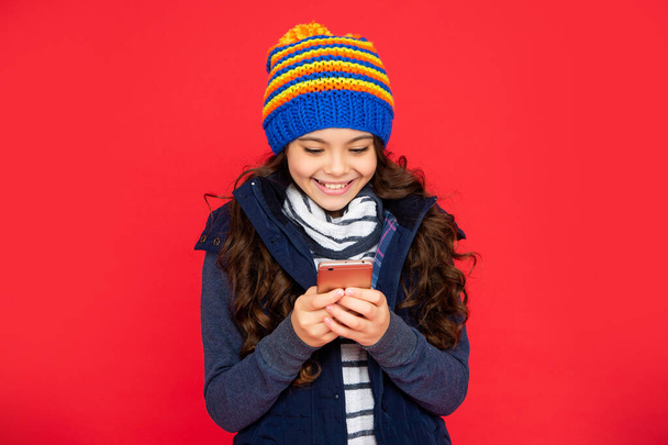 portrait of child blogging with phone. express positive emotion. greeting with winter holidays. happy kid chatting online in hat on red background. teen girl in down vest texting on smartphone. - Foto, afbeelding