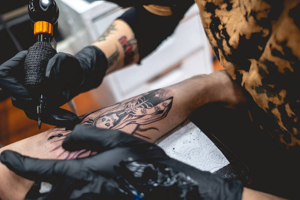 Detail van het jonge tattoo artiest meisje met bril en masker die een tattoo maakt van 'La Santa Muerte' (Onze Lieve Vrouw van de Heilige Dood) in de arm van een vrouw met tattoo machine - Foto, afbeelding