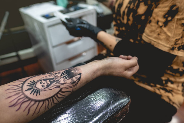 Деталь молодої дівчини-татуювальниці з окулярами і масками, яка виготовляє татуювання з "Богородиці Святої Смерті" в руці жінки з татуювальною машиною. - Фото, зображення