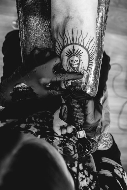 Detalle de joven artista del tatuaje con gafas y máscara haciendo un tatuaje de 'La Santa Muerte' en el brazo de una mujer con máquina de tatuaje (en blanco y negro)) - Foto, imagen