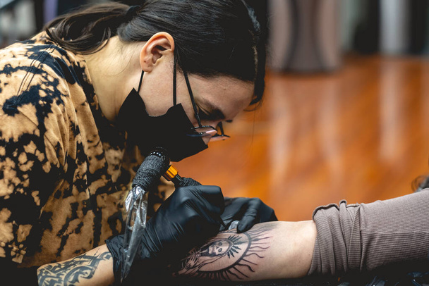 Részlet a fiatal tetováló művész lány szemüveggel és maszkkal, hogy egy tetoválás "La Santa Muerte" (Our Lady of Holy Death) a karjában egy nő tetováló gép - Fotó, kép