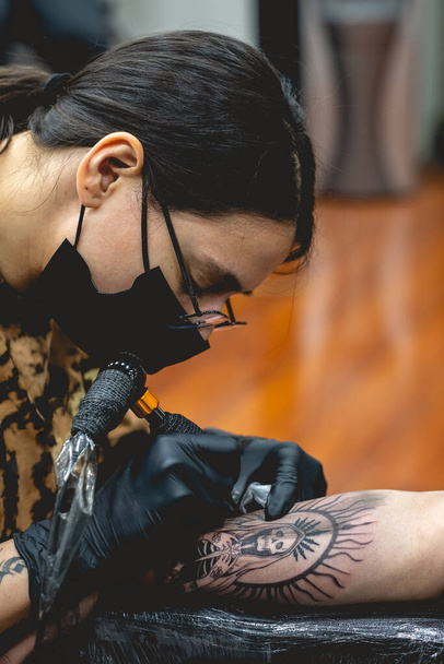 Detalle de joven artista del tatuaje con gafas y máscara haciendo un tatuaje de 'La Santa Muerte' en el brazo de una mujer con máquina de tatuaje - Foto, imagen
