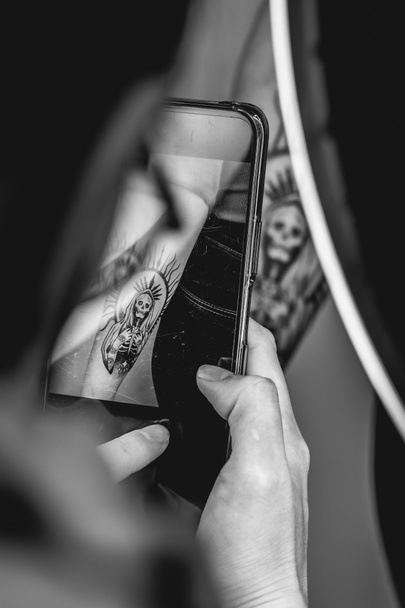 Detail of young tattoo artist girl with glasses and mask taking a photo with the phone of a new tattoo of 'La Santa Muerte' (Our Lady of Holy Death) in the arm of a woman (in black and white) - Foto, afbeelding