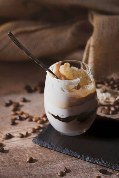 Glass of coffee milkshake with a metal spoon surrounded by coffee beans on an out of focus background. Selective focus. Coffee dessert concept. - Photo, Image