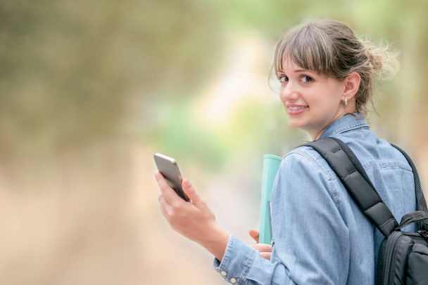 Улыбающаяся молодая женщина, держащая мобильный телефон, оглядываясь на камеру на фоне нефокусировки. Выборочный фокус. Технологическая концепция. - Фото, изображение