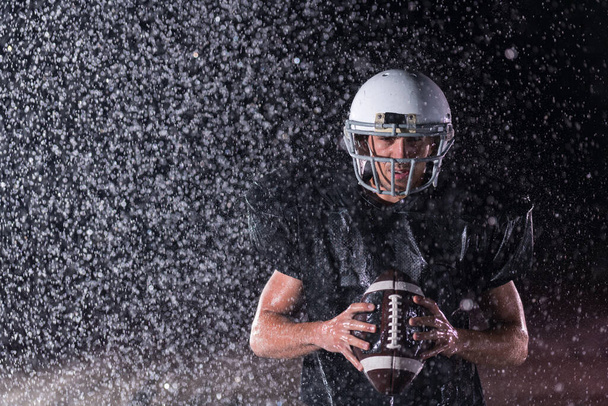 American Football Athlete Warrior Standing on a Field Holds his Helmet and Ready to Play. Player Preparing to Run, Attack and Score Touchdown. Rainy Night with Dramatic lens flare and rain drops. High - Zdjęcie, obraz