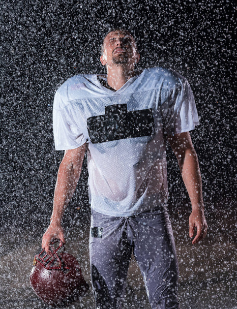 American Football Athlete Warrior Standing on a Field Holds his Helmet and Ready to Play. Player Preparing to Run, Attack and Score Touchdown. Rainy Night with Dramatic lens flare and rain drops. High - 写真・画像