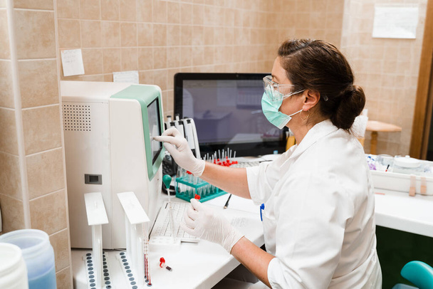 研究室のアシスタントは、血液分析器に取り組み、研究室で血液学的解析を行います。研究室の医療機器。血液の生化学的および血液学的解析 - 写真・画像