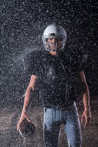 American Football Athlete Warrior Standing on a Field Holds his Helmet and Ready to Play. Player Preparing to Run, Attack and Score Touchdown. Rainy Night with Dramatic lens flare and rain drops. High - 写真・画像