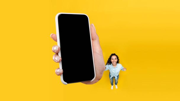 Wesoły arabski Kobieta Wyświetlanie Duży smartfon z pustym ekranem Polecam aplikację stojącą nad żółtym tle Studio. Świetna aplikacja mobilna. Panorama, makieta, widok z góry - Zdjęcie, obraz