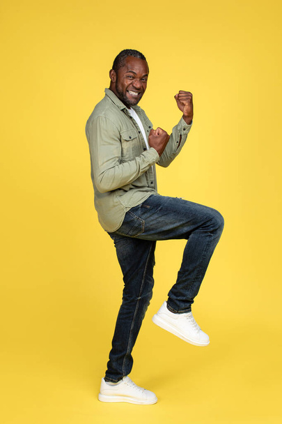 Satisfied happy middle aged african american man in casual rejoices to victory, celebrate success, makes win gesture, isolated on yellow background, full length, studio. People emotions and triumph - Foto, immagini