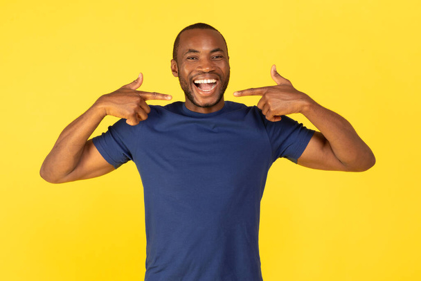 Portrét černocha ukazující prsty na jeho zubatý úsměv vyjadřující pozitivní emoce stojící nad žlutým pozadím v ateliéru, na sobě modré tričko. Podívej se na mě. - Fotografie, Obrázek
