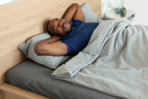 Afroamerykanin mężczyzna spania dobrze trzymając się za ręce za głowę drzemki pokryte kocem w przytulnym łóżku leży w nowoczesnej sypialni w domu. Koncepcja zdrowego snu - Zdjęcie, obraz