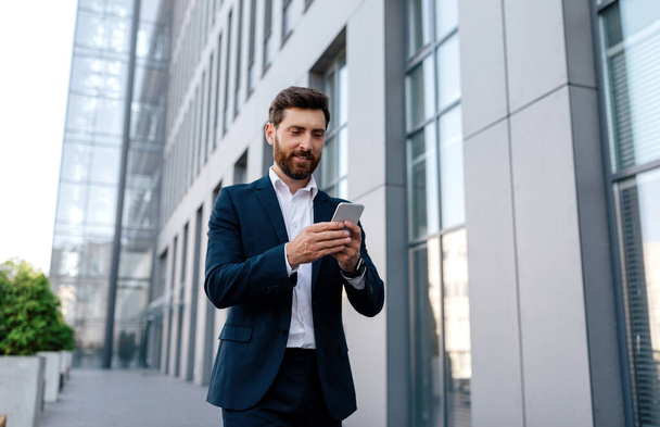 Busy glad confident attractive young caucasian man with beard in suit goes to work, has video call with smartphone outdoor near modern office building. Businessman and new app for business, new normal - Foto, immagini