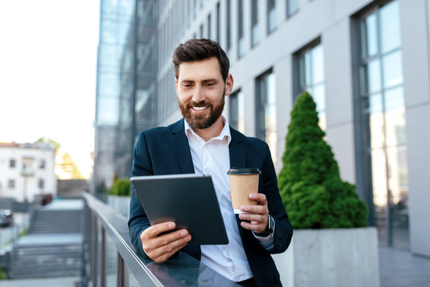 Smiling confident young caucasian guy with beard in suit has video call at tablet, hold cup of takeaway coffee outdoors on balcony of modern office building. Business, new normal for work with gadget - Фото, зображення
