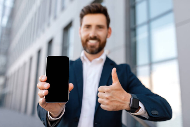 Glad confident young caucasian male with beard in suit shows smartphone with empty screen, shows thumb up near office building. Businessman advises app for trade, offer and business recommendation - Photo, image