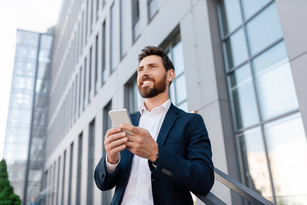 Cheerful confident young caucasian man with beard in suit typing on smartphone outdoor, looking at empty space near modern office building. Ad and offer for business, businessman use app for trade - Foto, imagen