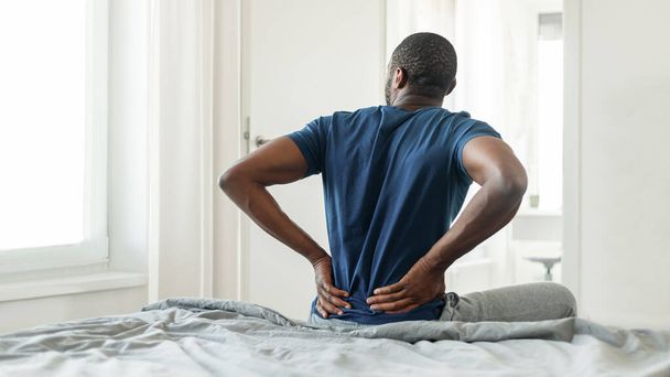 Takaisin Näkymä Afrikkalainen Amerikkalainen mies ottaa kipua koskettaa Alaselkä istuu sängyllä moderni makuuhuone sisätiloissa. Epämukava sänky ja selkärangan terveysongelma käsite. Panoraama - Valokuva, kuva