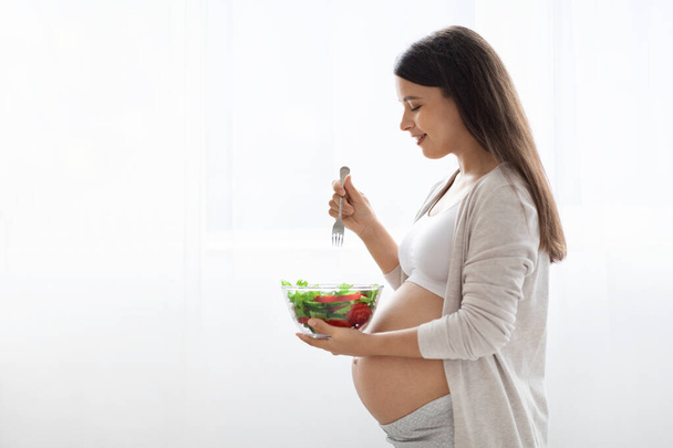 家の中で空腹の美しい長髪の若い妊娠中の女性は、ウィンドウの横に立って、健康的な新鮮な野菜のサラダを食べ、ビタミンやミネラルでいっぱいのおいしい食事を楽しんで、スペースをコピー - 写真・画像