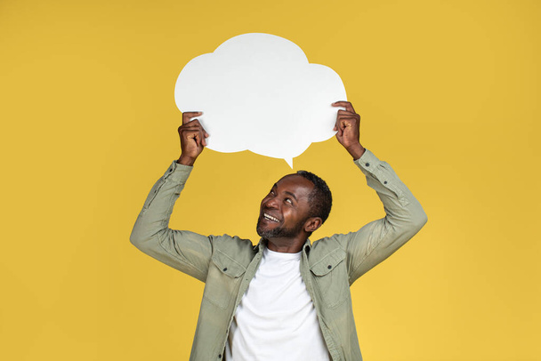 Cheerful handsome adult african american guy in casual looking at abstract cloud bubble with blank space for text, isolated on yellow background, studio. Ad and offer, thoughts and got idea, lifestyle - Foto, afbeelding
