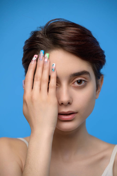 A skinny beautiful young brunette with her palm covering the right side of her face, at blue background - Фото, изображение