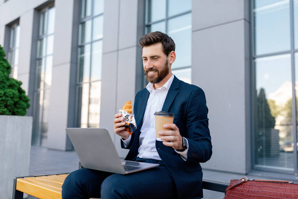 Successful business and lunch. Smiling handsome young caucasian man with beard in suit watches video on laptop, sits on bench with food and drink takeaway near office building. Coffee break and relax - Photo, Image