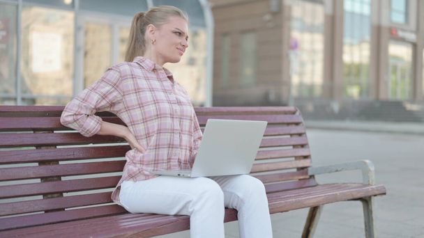 Donna casual con mal di schiena utilizzando il computer portatile mentre seduto all'aperto sul banco - Foto, immagini