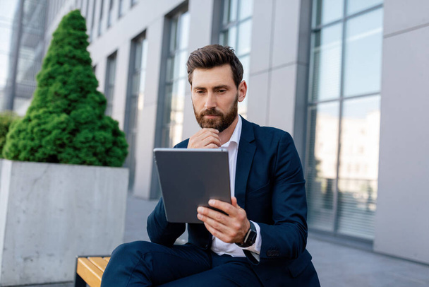 Concentrated pensive attractive young caucasian male with beard in suit reads news on tablet, sits on bench near modern office building. Businessman planning business strategy, new normal and app - Zdjęcie, obraz