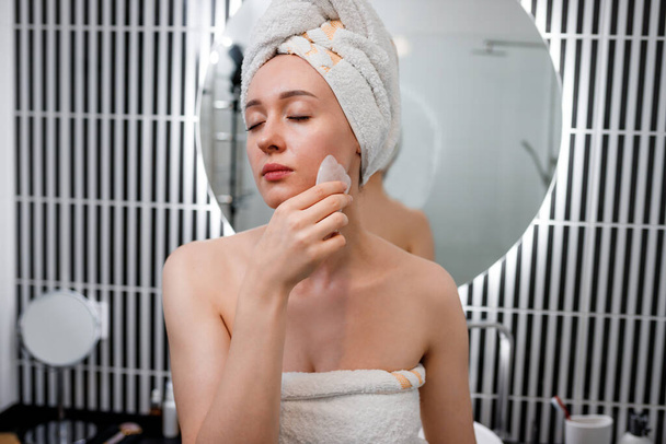 Young beautiful woman wearing white towel doing face massage with pink quartz scraper during beauty morning routine in bathroom. Anti aging treatments and skincare concept - 写真・画像