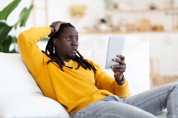 Megdöbbent afro-amerikai srác néz digitális tabletta képernyő otthon, zavarodott fiatal fekete férfi érzelmileg reagál a rossz hír, miközben ül a modern modul kanapén a nappaliban, szabad hely - Fotó, kép