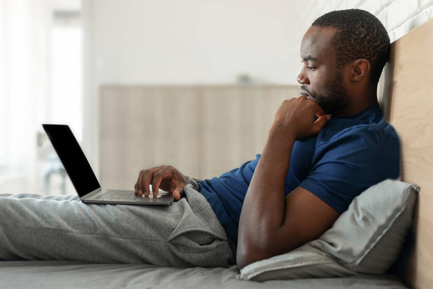 自宅のモダンなベッドルームでオンラインで作業を考えてベッドに座ってノートパソコンを使用して黒人男性のサイドビュー。フリーランスの男コンピュータ上でインターネットを閲覧します。距離の仕事と技術の概念 - 写真・画像