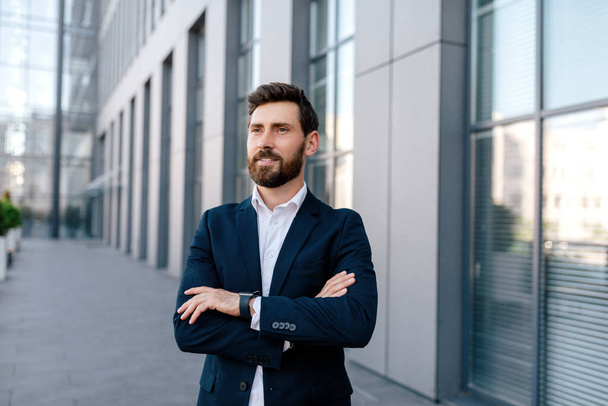 Serious glad confident attractive young caucasian male with beard in suit with crossed arms, looks at empty space near modern office building, outdoor. Businessman and startup, business and trade - Fotografie, Obrázek