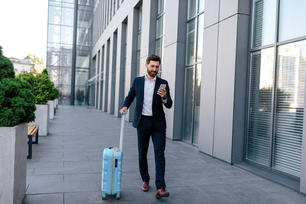 Happy confident handsome young caucasian male with beard in suit with suitcase goes, chatting by smartphone near modern office building, outdoor. Business trip, businessman use app map in gadget - Foto, imagen