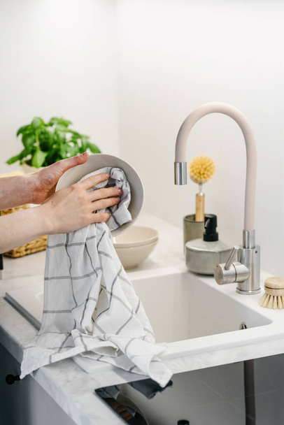 Gedeeltelijk uitzicht op een vrouw die borden veegt met handdoek in de keuken. Milieuvriendelijke vaatwasborstels bij het wassen van keramische borden. Thuis comfort. Huishoudelijke apparatuur. Huisvrouw plichten. Schoonmaakpersoneel - Foto, afbeelding