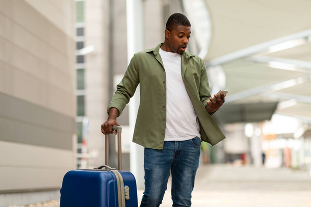 Шутливый чернокожий парень, читающий по телефону послание, стоит с чемоданом на улице аэропорта. Концепция проблем мобильной связи - Фото, изображение